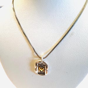 May include: Silver-toned necklace with a pendant featuring the words "LIVE FROM THE HEART" and a gold-colored heart. The pendant is rectangular with a textured border, suspended from a thin, silver chain.