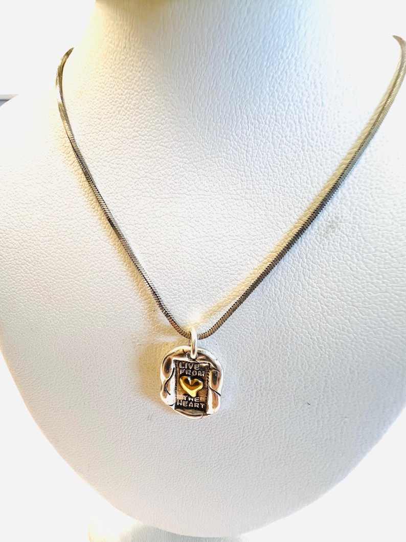 May include: Silver-toned necklace with a pendant featuring the words "LIVE FROM THE HEART" and a gold-colored heart. The pendant is rectangular with a textured border, suspended from a thin, silver chain.