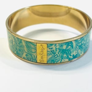 May include: Gold-tone bangle bracelet with a turquoise and white floral pattern. The bracelet features the text "Lilly Pulitzer" on a gold rectangular plate. The design includes flowers, leaves, and other decorative elements.