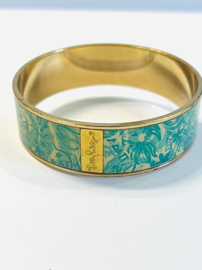 May include: Gold-tone bangle bracelet with a turquoise and white floral pattern. The bracelet features the text "Lilly Pulitzer" on a gold rectangular plate. The design includes flowers, leaves, and other decorative elements.