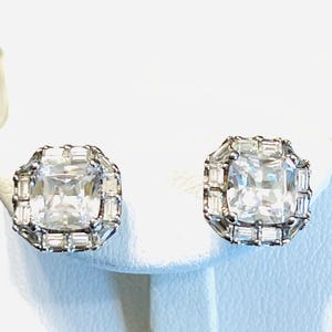May include: A pair of square-cut, clear gemstone stud earrings. Each earring features a large, square center stone surrounded by smaller, rectangular stones, set in a silver-toned metal. The earrings are displayed on a white surface.