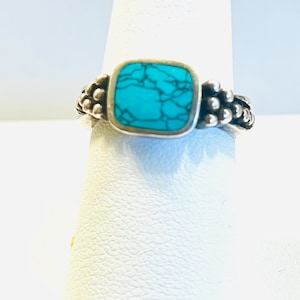 May include: A silver ring featuring a square, turquoise stone. The stone is set in a silver bezel and surrounded by a beaded design. The ring is displayed on a white background.