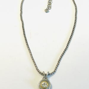 May include: A silver-toned necklace featuring a twisted rope chain and a circular pendant. The pendant has a central clear round gemstone surrounded by a gold-toned ring and a textured silver-toned border. It includes a lobster claw clasp.