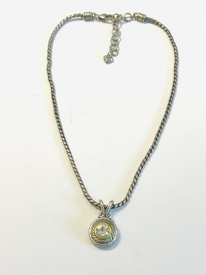 May include: A silver-toned necklace featuring a twisted rope chain and a circular pendant. The pendant has a central clear round gemstone surrounded by a gold-toned ring and a textured silver-toned border. It includes a lobster claw clasp.