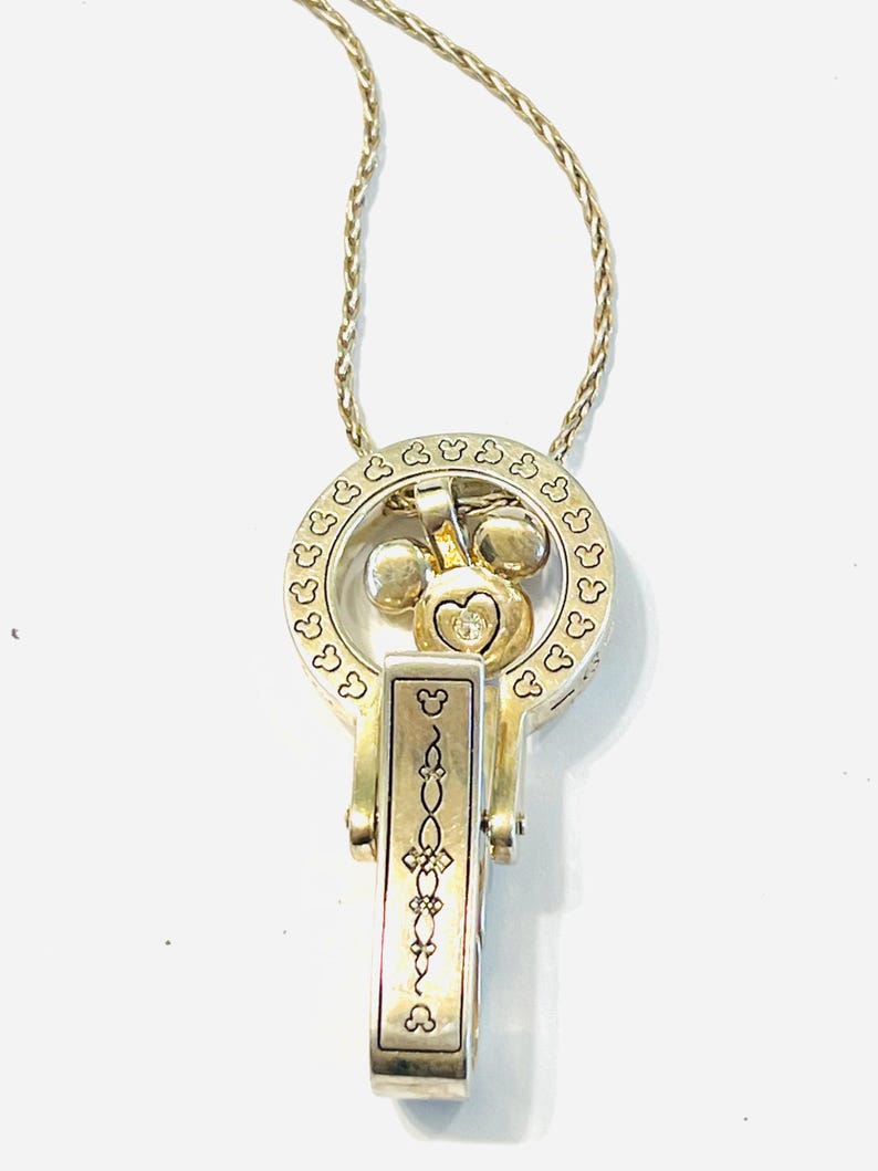 Puede incluir: Un collar plateado con un colgante tem&aacute;tico de Mickey Mouse. El colgante presenta un dise&ntilde;o circular con siluetas de la cabeza de Mickey Mouse, una cabeza de Mickey Mouse con una gema en forma de coraz&oacute;n y un dije rectangular con un patr&oacute;n adornado. La cadena es un dise&ntilde;o delicado y retorcido.