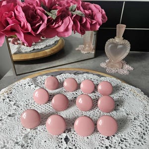 May include: A collection of pink, round cabinet knobs arranged on a white lace doily. A bouquet of pink flowers and a heart-shaped perfume bottle are in the background, along with a mirror.