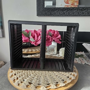 Puede incluir: Una caja de almacenamiento de diapositivas de fotos de plástico negro con múltiples ranuras. La caja se encuentra sobre una base redonda de madera con un tapete de ganchillo beige. Se ven flores rosas dentro.