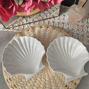 May include: Two white, shell-shaped dishes sit on a round, beige crocheted doily. The dishes have a glossy finish and are on a wooden tray. Pink flowers and a heart-shaped perfume bottle are in the background.