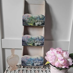 May include: A three-tiered, cream-coloured wooden wall shelf with three decorative compartments. Each compartment features a landscape painting with a house, trees, and colourful foliage. A pink heart-shaped perfume bottle and a vase of pink flowers are on the table.