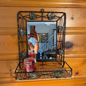 Peut inclure: Miroir rectangulaire encadré de métal noir avec une étagère en osier. Le cadre est orné de feuilles vertes et dorées. Une bougie rouge dans un pot en verre avec le texte "WINTER CANDY" est posée sur l'étagère.