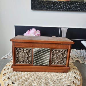 May include: A rectangular wooden box with a hinged lid, featuring two square side panels with an ornate, raised metallic scrollwork design and a central square panel covered in a fine, white and black checkered fabric mesh material.