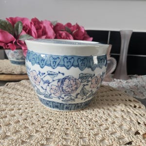 Peut inclure: Un bol en céramique avec un bord blanc et un motif floral bleu et rose. Le bol a une bordure décorative bleue et repose sur un napperon en crochet de couleur crème. L'arrière-plan comprend des fleurs roses et une surface noire et blanche.