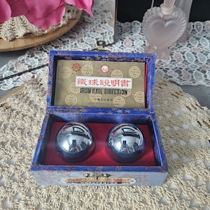 Peut inclure: Une boîte bleue avec un intérieur rouge contient deux boules métalliques argentées et brillantes. La boîte porte une étiquette dorée avec du texte chinois, indiquant "Iron Ball Direction". La boîte est posée sur une surface en crochet crème.