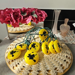 Può includere: Una stringa di luci gialle di Halloween con sagome di streghe nere. Le luci sono disposte su un vassoio di legno con un centrino beige all'uncinetto. Rose rosa e bottiglie di vetro decorative sullo sfondo.