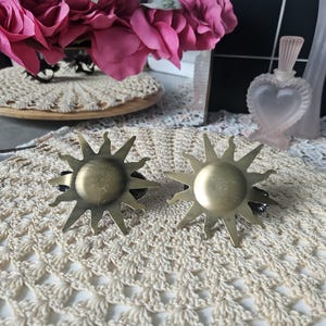 May include: Two circular brass-toned metal sunburst ornaments featuring a central disc surrounded by radiating pointed rays. Each sun-shaped piece has a brushed metallic finish and is attached to a dark base, presenting a celestial design.