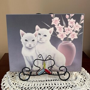 Peut inclure: Une impression encadrée représentant deux chatons blancs, un vase rose avec des fleurs et un présentoir décoratif en métal noir en forme de papillon avec des perles colorées. L'impression repose sur un napperon en crochet blanc.