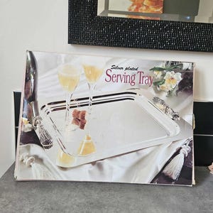 May include: A silver-plated serving tray with handles, shown on a box. The box depicts the tray with champagne flutes, a cork, and white tassels. The text "Silver plated Serving Tray" is displayed.