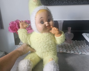 Könnte beinhalten: Eine Puppe mit blassem Gesicht, dunklen Wimpern und bemalten Gesichtszügen, bekleidet mit einem flauschigen, hellgelben Einteiler mit passender spitzer Mütze. Der Anzug hat einen weißen, strukturierten Besatz um Gesicht, Handgelenke und Füße sowie kleine hellgelbe Materialbüschel an den Spitzen der Mütze und der Füße.