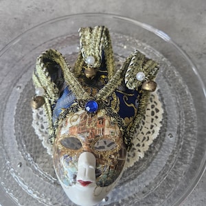 Pode incluir: Uma máscara veneziana decorativa com um rosto branco, lábios vermelhos e um design detalhado de paisagem urbana. A máscara apresenta detalhes em tecido dourado e azul, uma joia azul e pequenos sinos. Descansa sobre um prato de vidro transparente.
