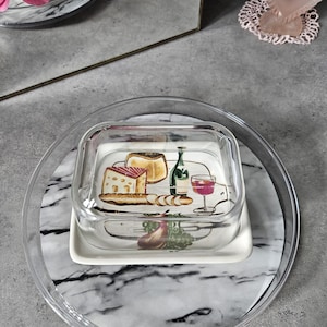 Peut inclure: Un plat à fromage en verre transparent avec une base blanche, orné d'un motif peint de fromage, de vin et d'un verre. Le plat repose sur un plateau à motifs de marbre, avec un couvercle en plastique transparent et un plateau de service en plastique transparent.