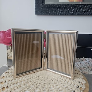 Peut inclure: Un cadre photo argenté à deux panneaux, ouvert et présenté sur une surface en bois ronde. Le cadre a un design simple avec une face en verre et un dos texturé beige. L'arrière-plan comprend un napperon décoratif et un miroir.