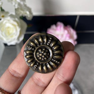 Peut inclure: Bouton de meuble rond en laiton antique avec un motif de tournesol. Le bouton présente un groupe central de petits points entourés de formes en forme de pétales. Le bouton est tenu dans une main, avec des bagues aux doigts.