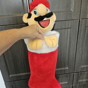 May include: A plush Christmas stocking featuring the character Mario, with a red hat, black moustache, and blue eyes. The stocking has a white faux fur cuff and a bright red velvet-like body, designed to hang from a red string loop at the top.