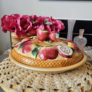 May include: A ceramic serving dish with a lid, featuring a basket weave design and a decorative apple motif. The lid is adorned with red apples and green leaves. The dish sits on a wooden base with a crocheted doily.