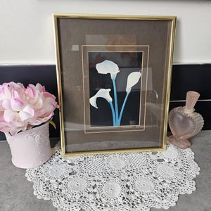 Peut inclure: Une œuvre encadrée représentant des callas blanches avec des tiges bleues sur fond noir. Le cadre doré repose sur un napperon en dentelle blanche. Un arrangement floral rose et un flacon de parfum en forme de cœur sont également visibles.