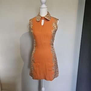 Peut inclure: Une robe courte orange sans manches avec un col et des panneaux latéraux à motifs floraux dorés et marron. La robe présente une ouverture en forme de goutte sous le col et une coupe ajustée, présentée sur un mannequin blanc.