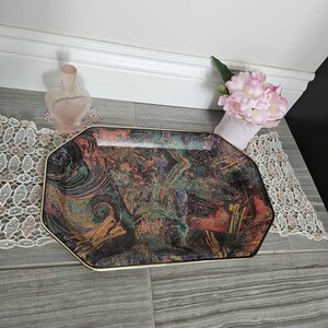 Puede incluir: Una bandeja de servicio octogonal con un borde dorado. La bandeja presenta un diseño abstracto colorido con remolinos de verde, naranja y morado. Una flor rosa y un frasco de perfume en forma de corazón están en el fondo.