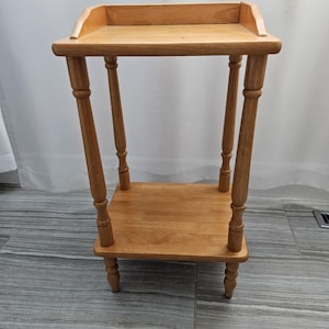 Könnte beinhalten: Kleiner, hellbrauner Beistelltisch aus Holz mit rechteckiger Oberseite und unterer Ablage. Der Tisch hat vier gedrechselte Beine und einen erhöhten Rand auf der Oberseite. Der Tisch ist etwa 61 cm hoch.