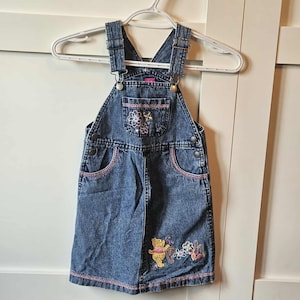 Peut inclure: Robe salopette en jean bleu avec des détails floraux brodés sur la poche de poitrine et un motif Winnie l'ourson sur la jupe. La robe a des coutures roses et des bretelles réglables, suspendue à un cintre blanc.