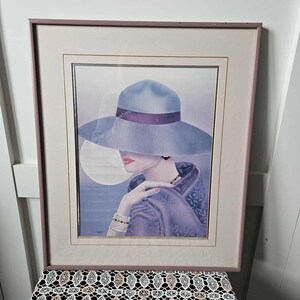 Peut inclure: Impression d'art encadrée représentant une femme portant un chapeau à larges bords et des vêtements violets. L'œuvre d'art a une esthétique vintage, avec des couleurs douces et une attention particulière au visage et aux accessoires du sujet. Le cadre est rose pâle.