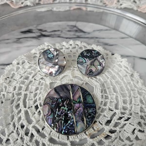Peut inclure: Ensemble de bijoux en coquille d'ormeau comprenant un broche ronde et des boucles d'oreilles assorties. La coquille d'ormeau irisée est sertie dans du métal argenté. La broche et les boucles d'oreilles sont présentées sur un napperon en crochet blanc.