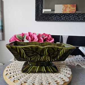 May include: A dark green ceramic centerpiece bowl with a leaf design, filled with pink artificial flowers. The bowl sits on a wooden base with a cream-coloured crocheted doily. A black beaded mirror is in the background.