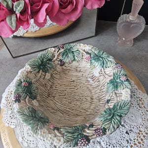 Può includere: Una ciotola decorativa in ceramica con un motivo a intreccio di cestino. Il bordo è decorato con foglie verdi, fiori bianchi e bacche viola. La ciotola poggia su un centrino di pizzo bianco e un tagliere di legno.