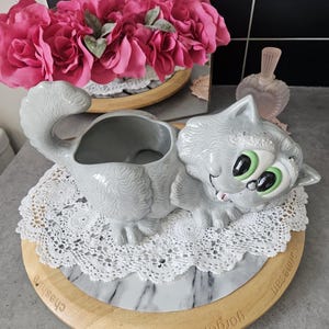 Peut inclure: Un pot de fleurs en céramique grise en forme de chat avec des yeux verts et une queue enroulée. Le pot est posé sur un napperon en dentelle blanche sur un plateau de service en bois et en marbre. Des fleurs roses sont en arrière-plan.