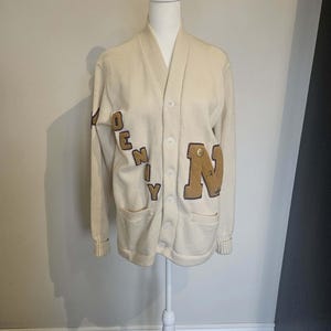 Peut inclure: Cardigan crème à col en V et fermeture boutonnée. Le pull porte les lettres "OENY" en marron, style universitaire, sur le devant. Deux poches sont visibles. Le pull est présenté sur un mannequin blanc.