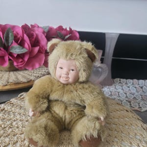 Könnte beinhalten: Eine Vintage-Puppe in einem Teddybär-Kostüm. Die Puppe hat ein Porzellangesicht mit rosigen Wangen und einem lächelnden Ausdruck. Das Bärenkostüm besteht aus weichem, braunem Kunstfell mit braunen Akzenten an Ohren und Füßen. Die Puppe sitzt auf einer gehäkelten Tischdecke.