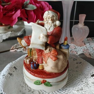 May include: A ceramic Santa Claus figurine, dressed in a red suit and hat, writing on a scroll. The figurine sits atop a round base with gifts and a sack. The base is white with a red top and green holly accents. A festive Christmas decoration.