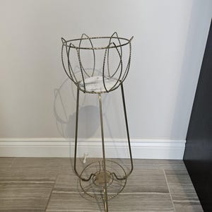 Puede incluir: Un soporte para plantas de estilo vintage con una estructura de metal dorado. El soporte presenta un diseño de cesta de alambre circular con una base con aspecto de mármol. Tiene tres patas y una base circular decorativa. Diseñado para una maceta.