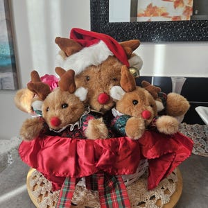 Peut inclure: Une décoration de Noël festive avec trois rennes en peluche dans un panier doublé de satin rouge. Les rennes ont une fourrure brune, des bois et des nez rouges. L'un porte un bonnet de Père Noël. Le panier est décoré d'un ruban à carreaux.