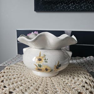 Peut inclure: Un cache-pot en céramique de couleur crème avec une finition mouchetée et un large bord ondulé. Le corps du cache-pot présente un motif floral peint de tournesols jaunes avec des feuilles et des tiges vertes, centré sur le devant du récipient.
