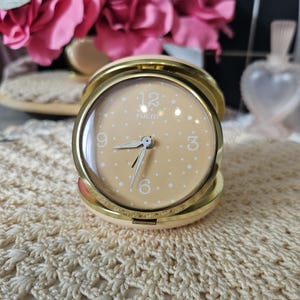 May include: A vintage, gold-toned travel alarm clock with a cream-colored face and white polka dots. The clock face displays the numbers 12, 3, 6, and 9. The clock is open, revealing the time. The word "EQUITY" is printed on the clock face.