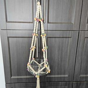 Puede incluir: Un colgador de plantas de macramé color crema con un cuenco de cristal transparente. El colgador está decorado con pequeños acentos florales de colores. El soporte para plantas está suspendido de un anillo de metal, diseñado para sostener una planta pequeña o un objeto decorativo.