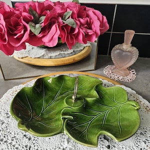 Peut inclure: Un plat de service vert en forme de feuille de céramique avec une poignée argentée. Le plat est posé sur un napperon en dentelle blanche. Des roses roses et un flacon de parfum en forme de cœur sont en arrière-plan.