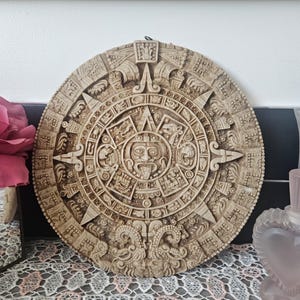 Peut inclure: Un calendrier aztèque circulaire beige, à accrocher au mur. La sculpture en relief détaillée présente un visage de soleil central entouré de symboles et de motifs complexes. C'est une pièce décorative.