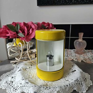 Peut inclure: Une lanterne vintage jaune et blanche avec une petite ampoule à l'intérieur. La lanterne est posée sur un napperon en dentelle blanche. Des roses roses et un miroir sont en arrière-plan.