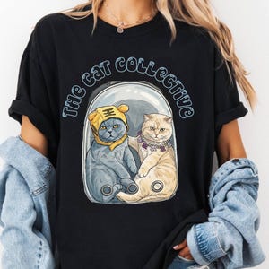 May include: Black t-shirt with a graphic of two cats inside a clear capsule. One cat wears a yellow hat, the other a purple necklace. The text "The Cat Collective" arches above the cats in light blue.